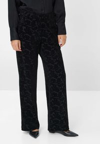 Schwarze Samt-Hosen mit weitem Bein, die ein strukturiertes Blumenmuster aufweisen, einen elastischen Bund haben und mit eleganten schwarzen spitz zulaufenden Ballerinas kombiniert sind.