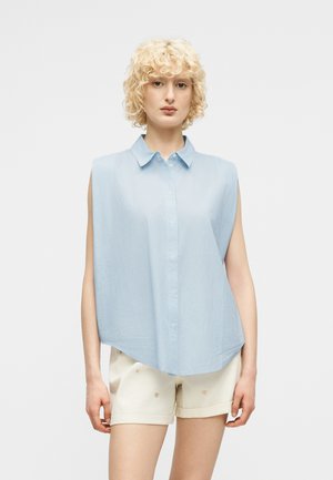 Kobieta o krótkich kręconych blond włosach, ubrana w jasnoniebieską bezrękawnikową koszulę na guziki i beżowe szorty z drobnymi wzorami.