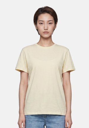 Jeune femme aux cheveux courts et foncés, portant un t-shirt jaune clair uni et un jean bleu, debout devant un fond neutre.