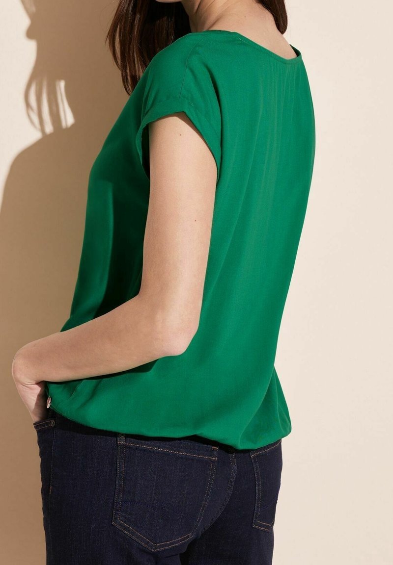 Femme portant un chemisier vert à manches courtes et un jean bleu foncé, debout avec une main dans la poche sur un fond beige.