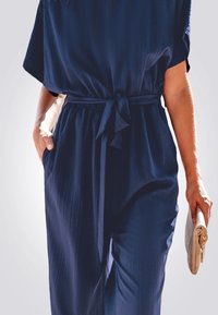 Une combinaison bleu marine à coupe ample, avec des manches courtes et une taille cintrée. Elle présente des rayures verticales et des poches latérales. Elle tient un sac à main de couleur claire.