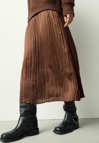 Person trägt einen braunen Faltenrock, ein braunes langärmeliges Oberteil und schwarze kniehohe Lederstiefel mit Schnallendetail und steht vor einer schlichten Wand.