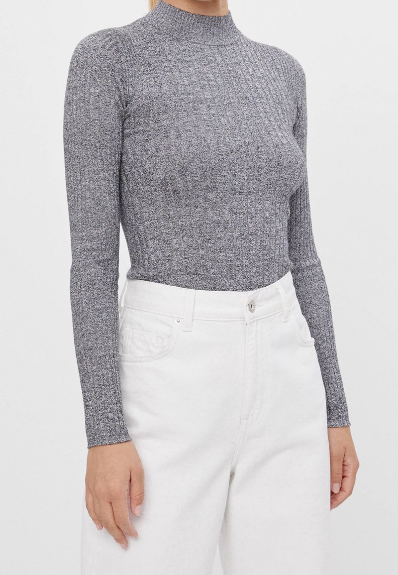 Femme portant un pull gris à col roulé côtelé ajusté à manches longues et un jean blanc taille haute avec poches avant.