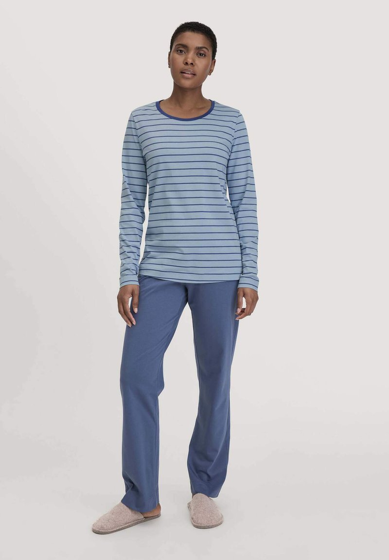 Langärmliges blau gestreiftes Oberteil mit einem Rundhalsausschnitt, kombiniert mit einfarbigen blauen Hosen. Das Model trägt weiche Hausschuhe und präsentiert ein entspanntes Outfit.