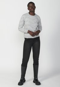 Maglione grigio lavorato a maglia con motivo a zigzag e piccoli fori, abbinato a jeans skinny neri e stivali neri alti fino al ginocchio.