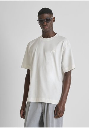 Jonge man met een oversized wit T-shirt, lichtgrijze broek en grote donkere zonnebril, staand tegen een effen lichte achtergrond.