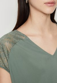 Top verde con scollatura a V, tessuto morbido e dettagli in pizzo sulle spalle, che creano un contrasto testurizzato rispetto al materiale liscio.