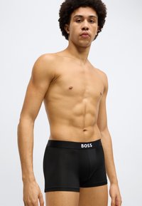 Fekete boxerrövidnadrág sima textúrával, széles derékpánttal, amelyen fehér "BOSS" logó található. Testhezálló szabás, minimalista stílus.