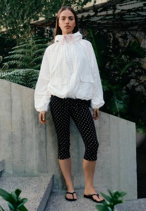 Kvinde, der står udendørs iført en hvid hættetrøje og sorte kortere leggings med hvide prikker, kombineret med sorte sandaler.