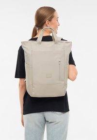 Beige hátizsák, dupla felső fogantyúval, oldalsó zsebbel és elülső cipzáras zsebbel, tartós anyagból készült. Egyszerű, strukturált dizájn logó részlettel.