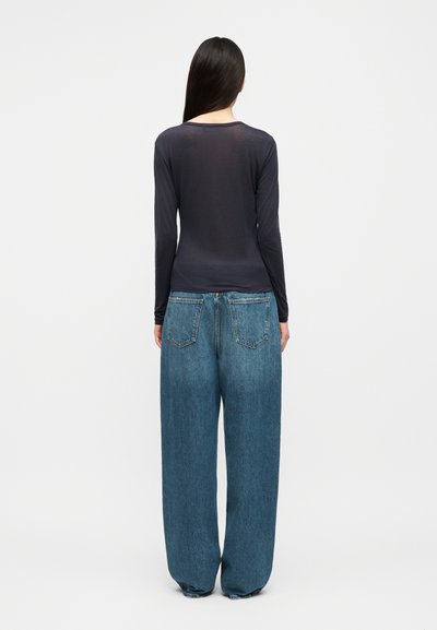 Sieviete ar gariem tumšiem matiem, valkājot pieguļošu melnu krekliņu ar garām piedurknēm un vaļīgas, platām kāju bikšu zilas džinsas, stāvot ar muguru pret skatītāju uz vienkārša fona.