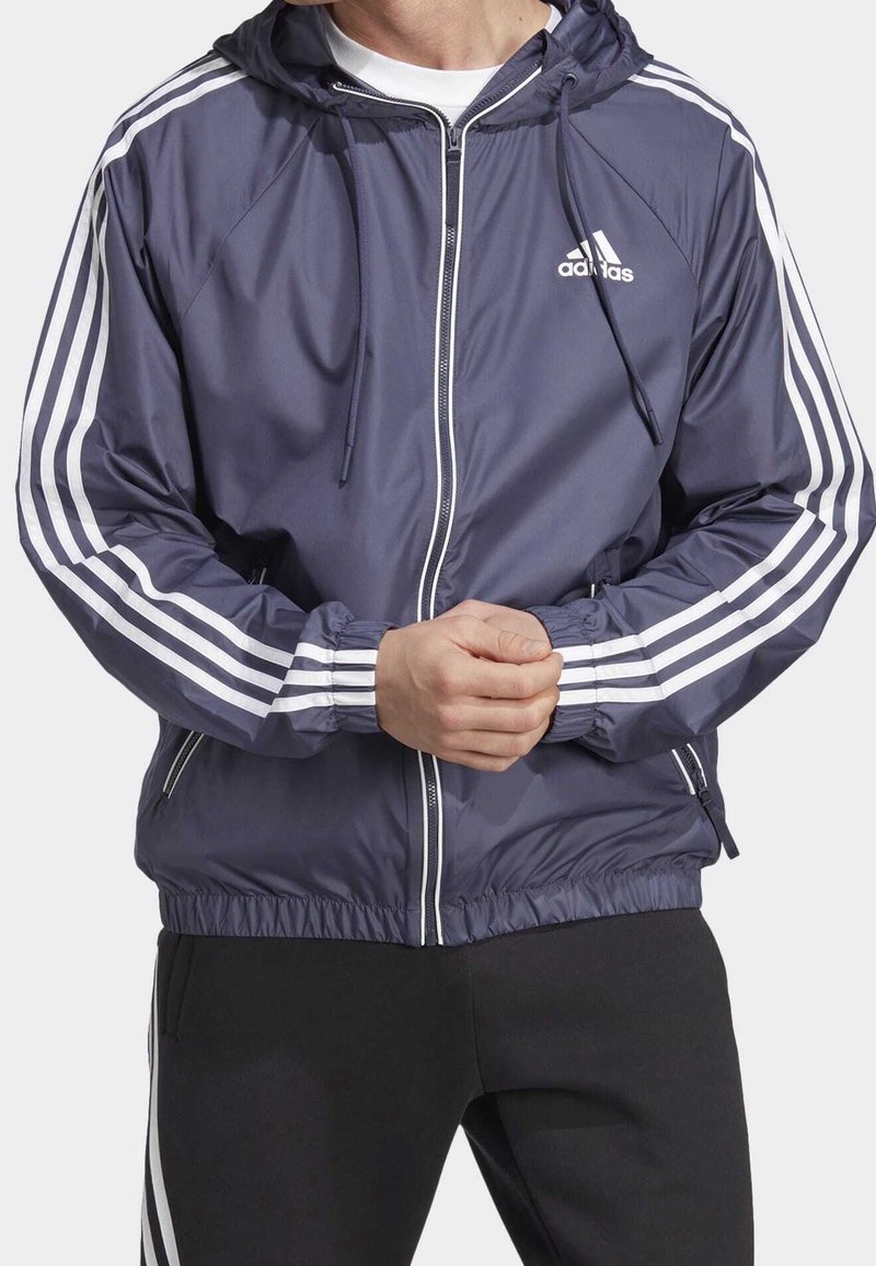 Homme portant une veste zippée Adidas bleu marine avec des bandes blanches sur les manches et un pantalon noir avec des bandes blanches sur les côtés. Mains jointes devant.