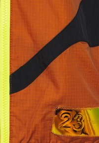 Orangekappa av tyg med rutmönster, svarta detaljer och en glansig guldpin som har siffran 23. Gul dragkedja. Texturerad yta.