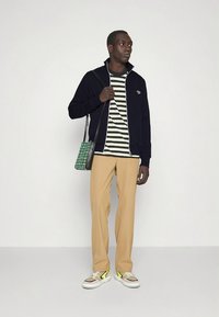 Námornícka bunda na zips cez zeleno-bielu pruhovanú košeľu, béžové nohavice a viacfarebné tenisky. Zelená károvaná taška visí z ramena.