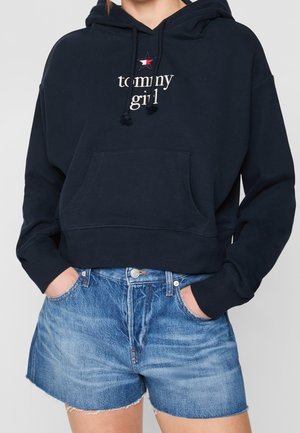 Kvinde iført en sort hættetrøje med "tommy girl" tekst og stjerne logo, kombineret med blå denimshorts, hænderne i lommerne.