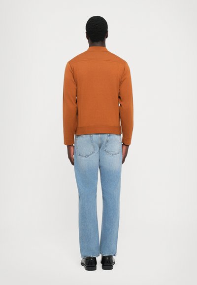 Ruskea puuvillatakki suoran leikkauksen omaavana, yhdistettynä vaaleansinisiin suoriin farkkuihin. Sileä pinta, minimalistinen muotoilu ja ilman näkyviä yksityiskohtia.