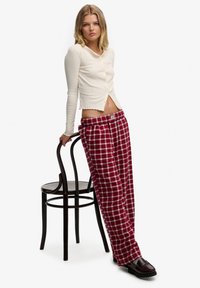 Kremowy, prążkowany top z długim rękawem, falistym wykończeniem na dole i guzikami, w połączeniu z luźnymi czerwonymi spodniami w kratę oraz ciemnymi mokasynami, stojący przy krześle.