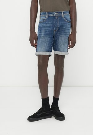 Közép kék színű farmer rövidnadrág fakó hatással, hajtott szegélyekkel és klasszikus öt zsebes kialakítással, fekete tornacipővel és zoknival párosítva.