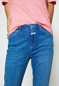 Personne portant un jean en denim bleu avec fermeture éclair et bouton à l'avant, associé à un t-shirt rose clair, les mains reposant derrière le dos.