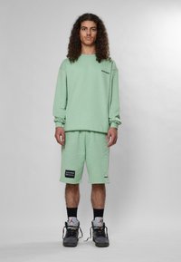 Felpa verde menta con maniche lunghe e collo tondo, abbinata a pantaloni corti verde menta. Pannello con logo nero sui pantaloni, scarpe da ginnastica grigie.