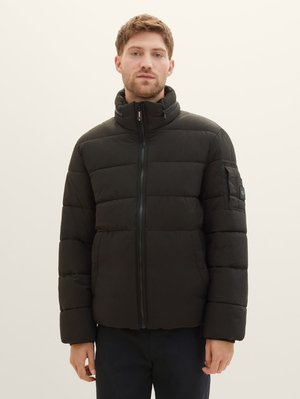 Chaqueta acolchada negra con cuello alto, cierre de cremallera y diseño acolchado. Cuenta con un bolsillo con cremallera en la manga izquierda. Textura suave.