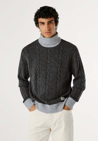 Maglione a coste in grigio scuro con colletto a imbuto e polsini in grigio chiaro, caratterizzato da un motivo testurizzato e etichette sull'orlo.