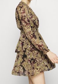 Blumenkleid mit langen Ärmeln, Peplum-Taille und schichted Rock. Der Stoff zeigt grüne, lila und braune Farbtöne in einem strukturierten Muster.