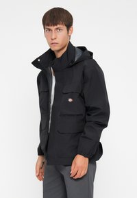 Black waterproof jacket with multiple front pockets, elastic cuffs, and a hood. Features a logo on the chest and a zip closure.