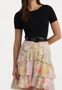 Černý žebrovaný top s krátkým rukávem, doplněný o vícevrstvovou sukni s pastelovými květinovými vzory v růžové, žluté a zelené barvě. Součástí je také lesklý černý opasek.