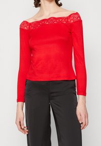 Röd långärmad topp med off-the-shoulder-design, spetsdetalj längs halsringningen och en figurnära silhuett. Kombinerad med mörka byxor.