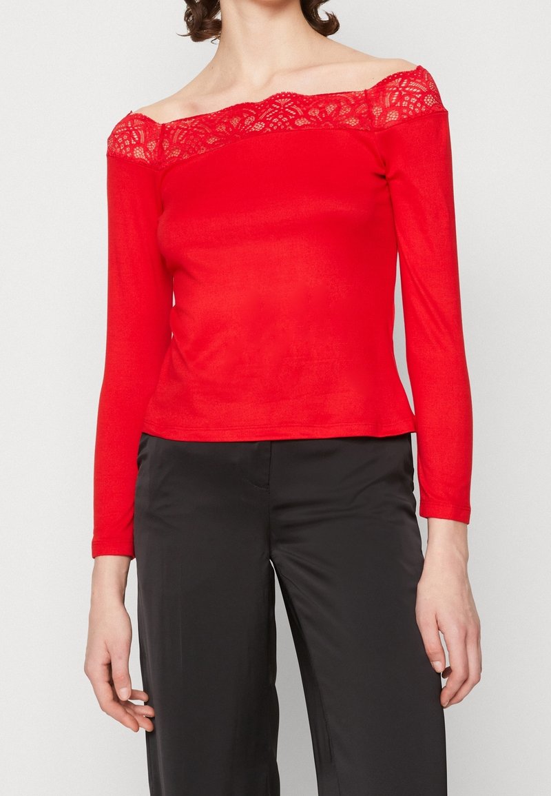 Röd långärmad topp med off-the-shoulder-design, spetsdetalj längs halsringningen och en figurnära silhuett. Kombinerad med mörka byxor.