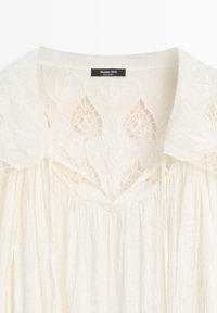 Blusa color crema con ampio colletto e dettagli floreali in pizzo sulla parte superiore del petto e sulle spalle, etichettata Massimo Dutti.
