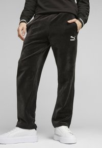 Pantalones de chándal de pana negra con cinturilla elástica, bolsillos laterales y un logo blanco de Puma impreso en el muslo izquierdo. Combinados con zapatillas blancas.