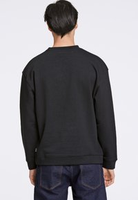 Sudadera negra con una textura suave, cuello redondo y hombros caídos. Llevada con jeans oscuros de mezclilla. Diseño minimalista, sin patrones.