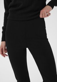 Leggings neri realizzati in materiale elastico, con una texture liscia e una silhouette aderente. Mano posizionata sulla vita per supporto.