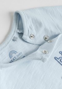 Body bébé bleu clair avec boutons-pression le long de l'épaule et de l'encolure, présentant un petit motif brodé et une étiquette "NEXT baby" à l'intérieur.