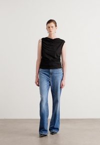 Svart ermeløs topp med drapert halslinning, parret med lyseblå flare jeans. Modellen står mot en nøytral bakgrunn.