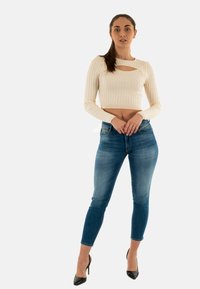 Top corto a maniche lunghe a coste color crema con dettaglio a taglio, abbinato a jeans skinny blu a vita media e tacchi alti neri.