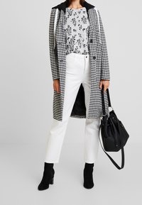 Czarny i biały płaszcz w kratę houndstooth z czarnym kapturem, szara bluzka w kwiaty, białe jeansy o prostym kroju oraz czarne botki na obcasie, trzymając czarną torebkę.