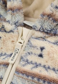 Nærbillede af en mønstret fleecejakke i beige, blå og brun med lynlås, der har et stofmærke med teksten "HUGS."