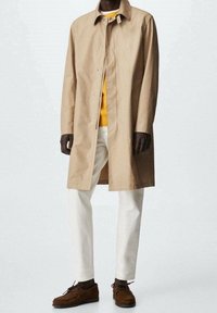 Trench coat beige con colletto, abbinato a un maglione giallo, pantaloni bianchi e scarpe in suede marrone. Tessuto morbido e silhouette dritta.