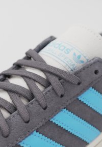 Grå mockasneaker med blå läderremsor, vit tunga med branding och grå snören. Har texturerad ovandel och kontrasterande sömmar.