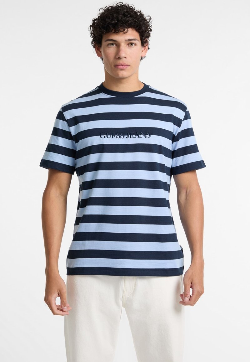 T-shirt rayé en bleu et marine, fabriqué en coton, avec des manches courtes et un col rond, portant le logo "GUESS JEANS" sur la poitrine.