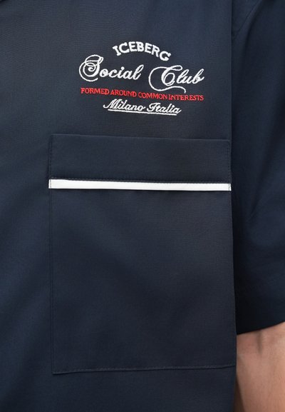 Chemise à manches courtes bleu marine avec une poche poitrine. Présente des passepoils blancs et une broderie complexe blanche et rouge avec le texte "Iceberg Social Club".