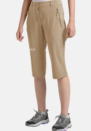 Beige knielange Outdoor-Shorts mit Reißverschlusstaschen, getragen von einer Person mit grauen Wanderschuhen und weißen Socken auf einfarbigem Hintergrund.