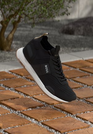 Schwarzer BOSS-Sneaker mit weißer Sohle, der über einem strukturierten Holz- und Steinboden schwebt, im Hintergrund unscharfer Baum und Wand.