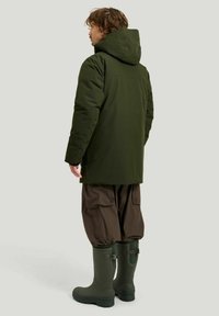 Grön isolerad jacka med huva, ihopparad med bruna cargobyxor och gröna gummistövlar. Slät textur, minimalistisk design.