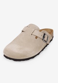 Zuecos de ante beige con punta redondeada, una correa de cuero y una hebilla de metal negra; superficie texturizada y plantilla de corcho. Suela de goma negra.