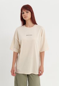 T-shirt oversize beige en tissu doux, avec des manches courtes et un col rond. Texte "POSITIF SINCÈRE" imprimé au centre.