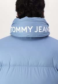 Giacca puffer azzurra chiara con colletto alto e la scritta "TOMMY JEANS" in bianco in grassetto. Tessuto liscio con texture gonfia.